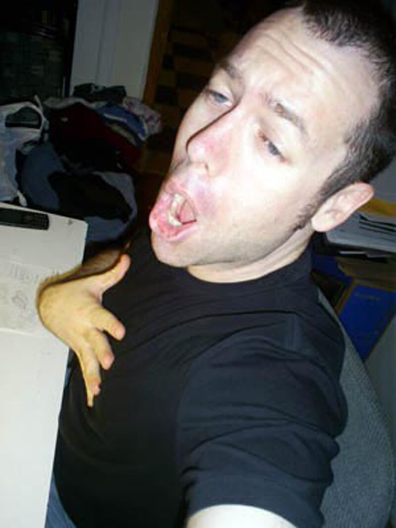 high-angle selfie of a man wearing a black t-shirt with mouth wide open while singing, hand on his chest.