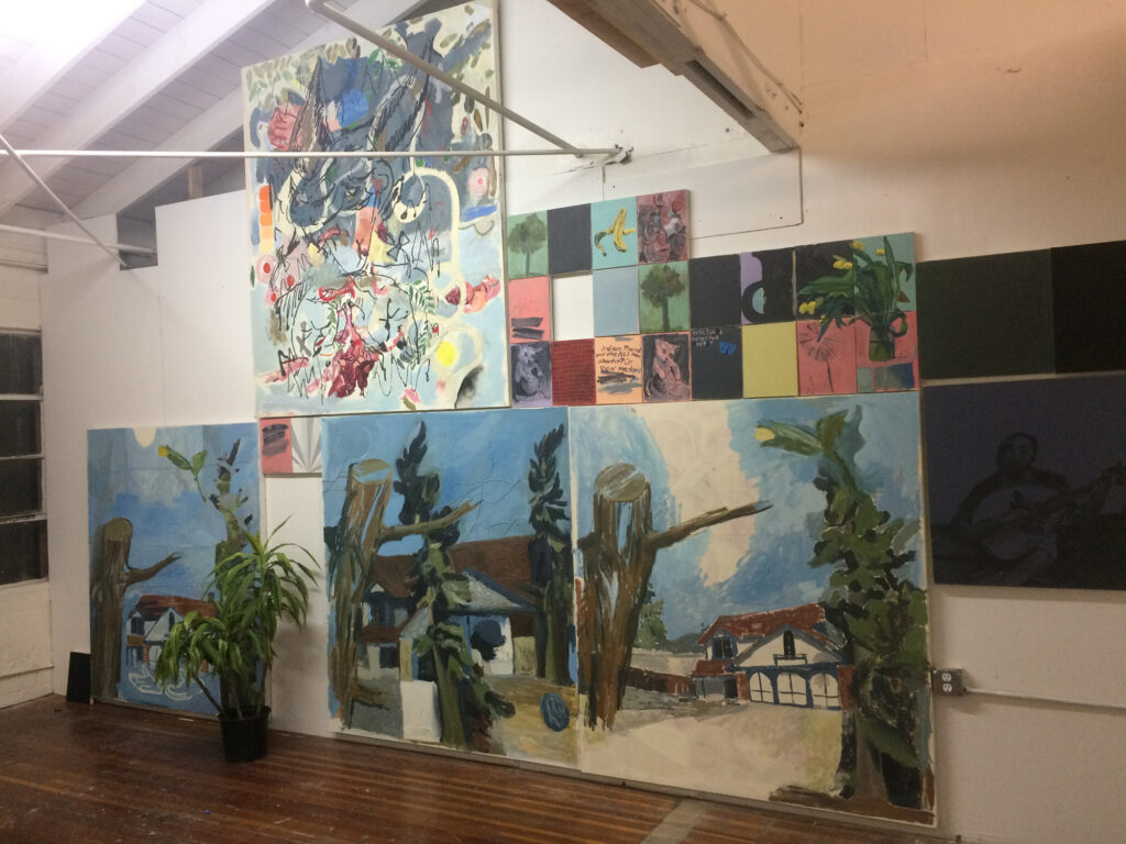 Art studio wall featuring paintings hung side-by-side from floor to ceiling with a potted plant in the center. 